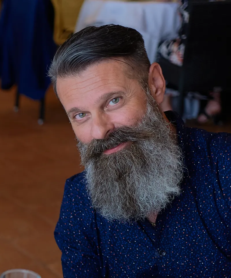 Ritratto di Giuseppe Irranca con la camicia blu, la barba lunga e un sorriso a testa inclinata